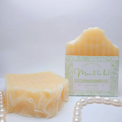 2 full-size bars of cream-colored soap one has a light green label printed with "Meant to Be! John & Annie, August 15, 2023." They are on a white background with a string of pearls as an accent. 
