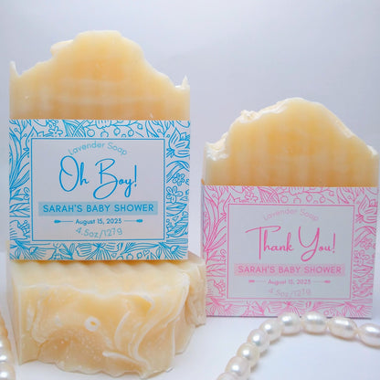 3 bars of cream-colored soap 1 has a pink label printed with "Thank You! Sarah's Baby Shower, August 15, 2023" and 1 blue label with "Oh Boy! Sarah's Baby Shower, August 15, 2023." They are on a white background with a string of pearls as an accent.