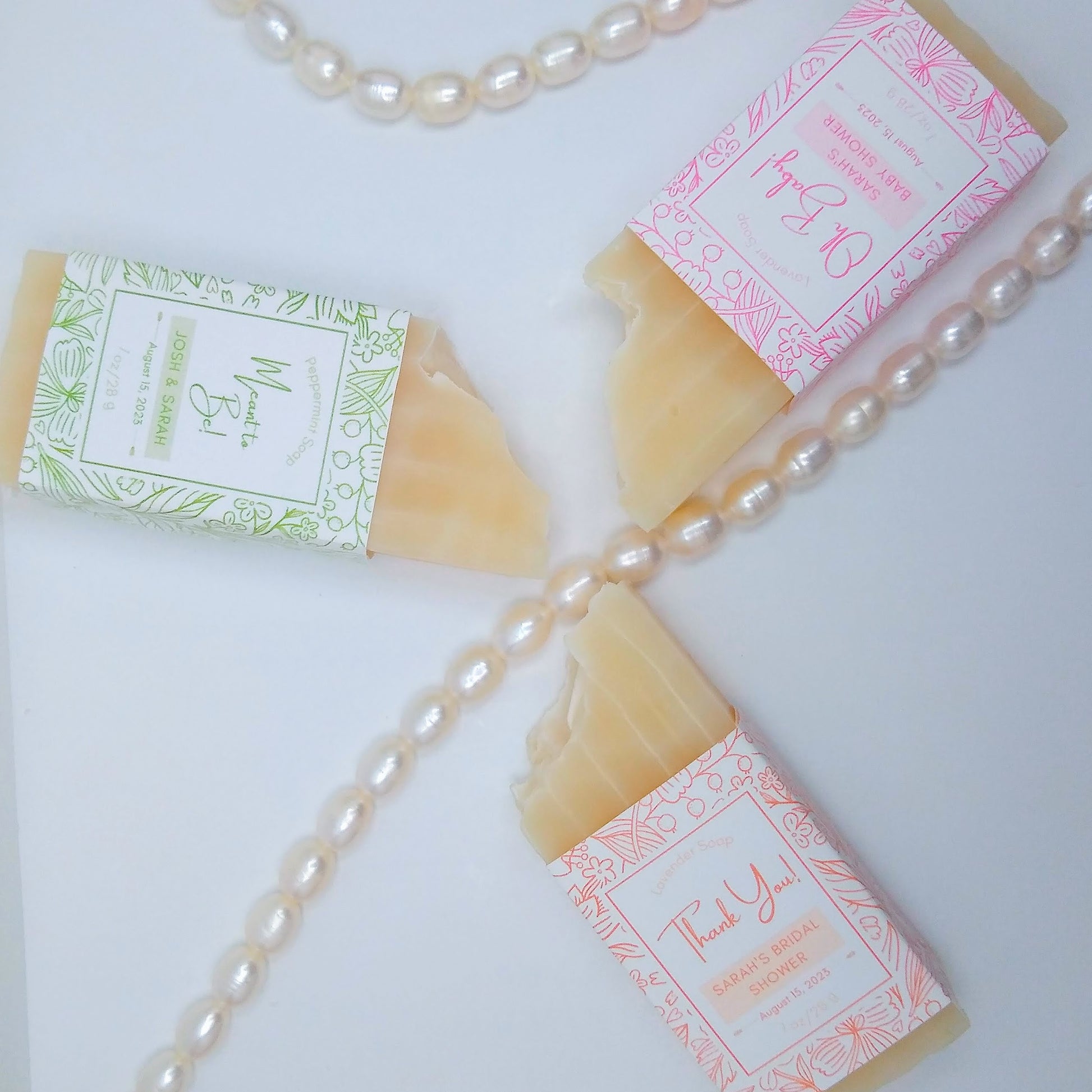 3 mini bars of cream-colored soap in a pinwheel shape around a string of pearls. One has a light green label, another an orange label, and another a pink label.
