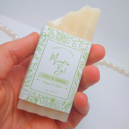 A mini bar of cream-colored soap held in a hand with a light green label printed with Meant to Be! John & Annie, August 15, 2023. They are on a white background with a string of pearls as an accent.