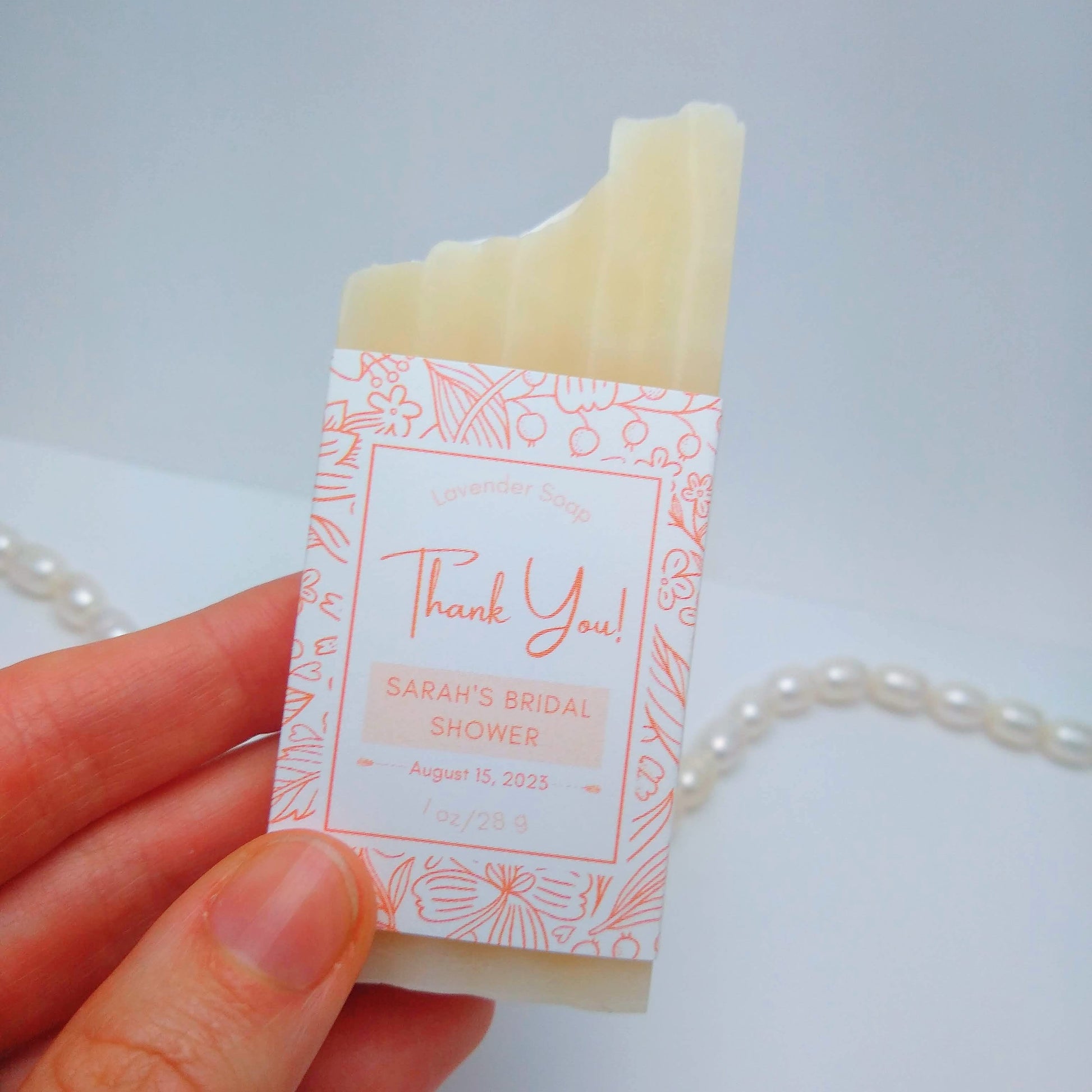 A mini bar of cream-colored soap held in a hand with a light orange label printed with Thank You! Sarah's Bridal Shower, August 15, 2023. They are on a white background with a string of pearls as an accent.
