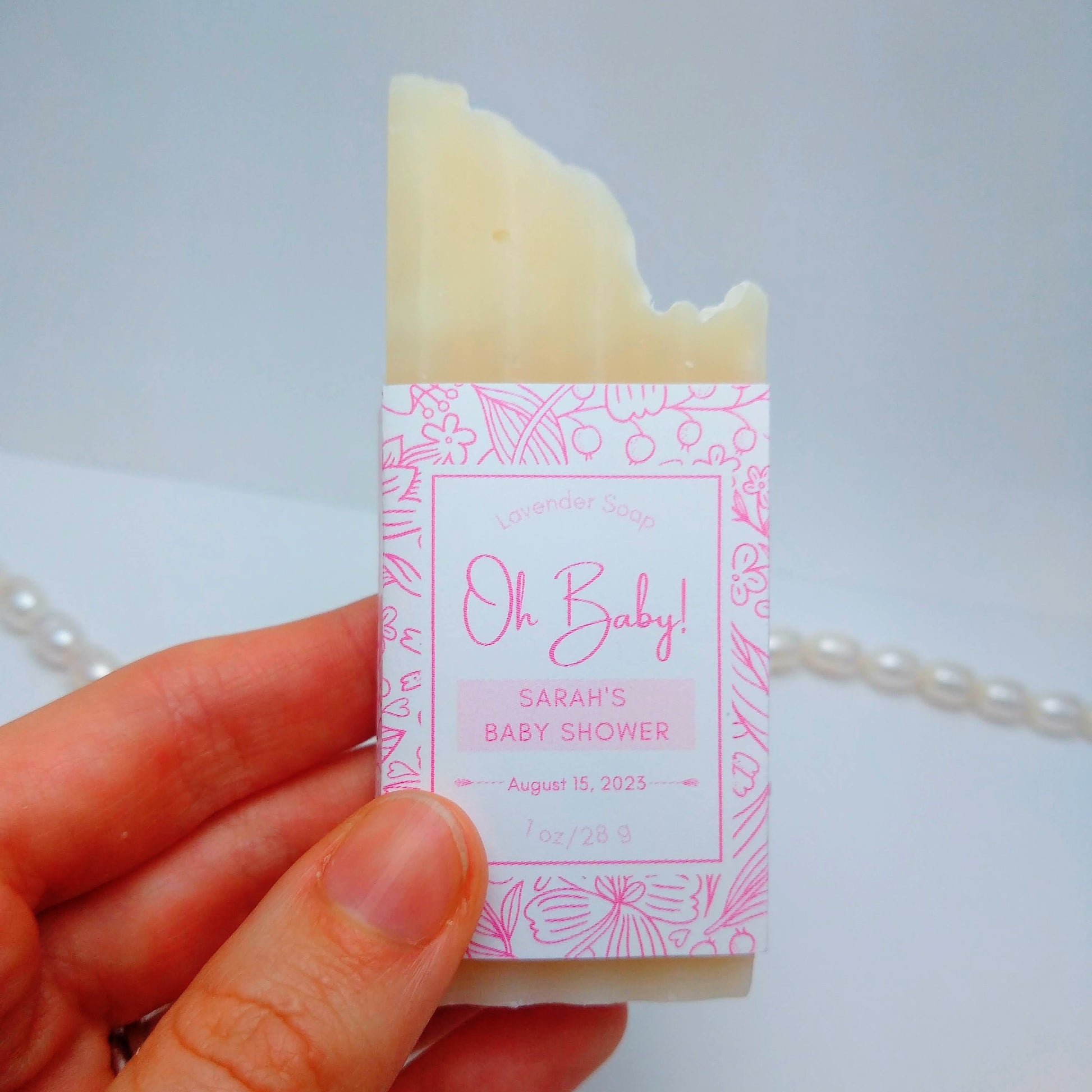A mini bar of cream-colored soap is held in a hand with a string of pearls in the background. The soap has a pink label printed with Oh Baby!, Sarah's Baby Shower, August 15, 2023. 